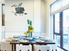 Moderní nástěnné hodiny ve tvaru šálku kávy s nápisem 'coffee time' a zrnky kávy, v tmavě zelené barvě, visí v elegantním interiéru nad dřevěnou konzolí s vázou a svíčkou Papilora.