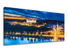 Na plátně je zobrazeno noční panorama Bratislavy s osvětleným hradem a mostem přes Dunaj, pod tmavě modrou oblohou s oranžovými odlesky západu slunce, Papilora.