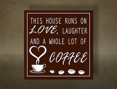 Hnědý obraz s bílým nápisem „THIS HOUSE RUNS ON LOVE, LAUGHTER AND A WHOLE LOT OF COFFEE“ a šálkem kávy s kávovými zrny, viditelný na dřevěném rámu Papilora.