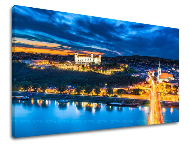 Obraz na plátně zachycuje noční panorama Bratislavy s osvětleným hradem a mostem přes Dunaj v modrých a oranžových tónech, Papilora.