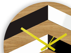 Moderní nástěnné hodiny s geometrickým vzorem v černé, bílé a dřevěné barvě se žlutými ručičkami visí v elegantním interiéru nad dřevěnou konzolí s vázou a svíčkou Papilora.