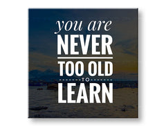 Obraz s citátem „You are never too old to learn“ visící v moderním interiéru nad dřevěnou konzolí s vázou a svíčkou Papilora.