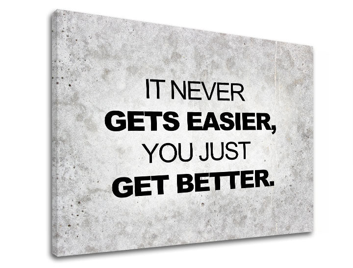 Plátno s motivačním citátem „IT NEVER GETS EASIER, YOU JUST GET BETTER.“ na šedém texturovaném pozadí, s viditelným dřevěným rámem, Papilora.
