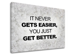 Plátno s motivačním citátem „IT NEVER GETS EASIER, YOU JUST GET BETTER.“ na šedém texturovaném pozadí, s viditelným dřevěným rámem, Papilora.