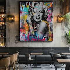 Obraz Marilyn Monroe v pop-art stylu s barevnými graffiti a zlatými detaily visí v moderním interiéru nad dřevěnou konzolí s vázou a svíčkou Papilora.