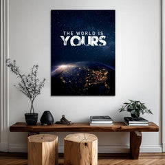 Obraz s motivem noční Země z vesmíru a bílým textem 'THE WORLD IS YOURS' visí v moderním interiéru nad dřevěnou konzolí s vázou a svíčkou Papilora.