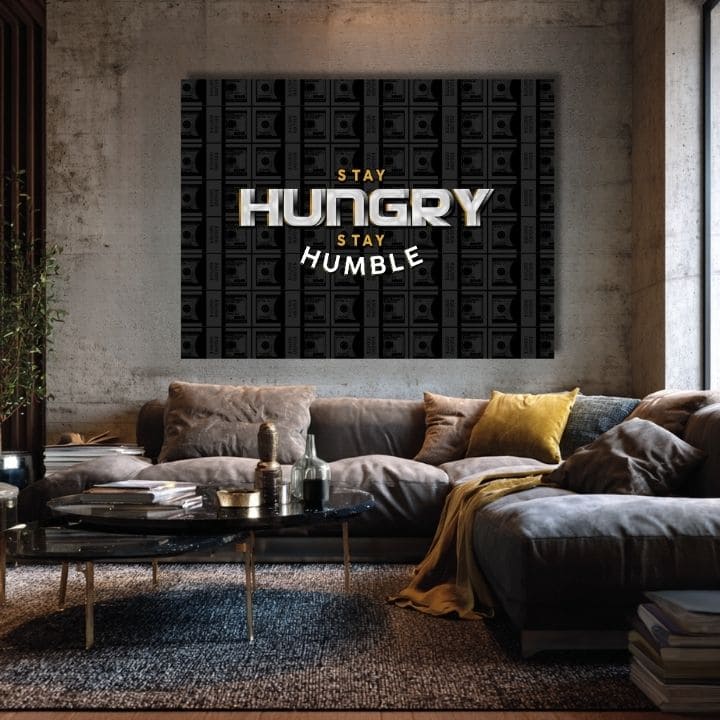 Obraz s motivačním citátem „STAY HUNGRY STAY HUMBLE“ a pozadím bankovek visí v moderním interiéru nad dřevěnou konzolí s vázou a svíčkou Papilora.