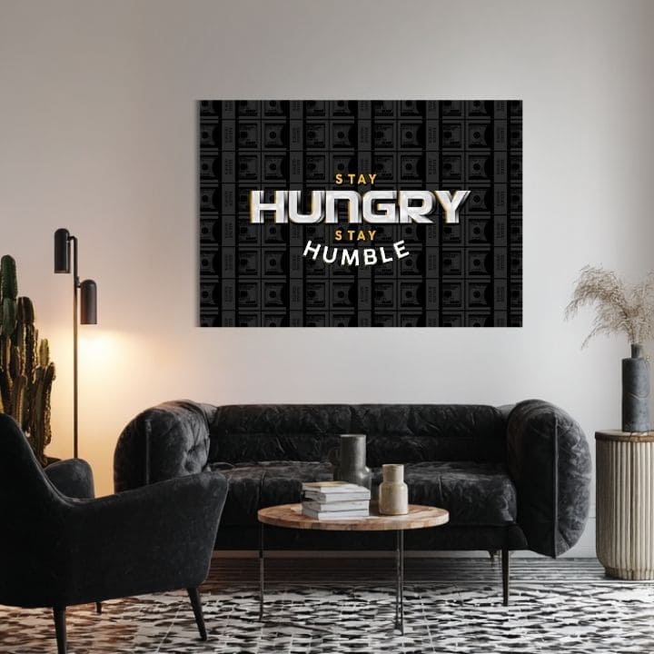 Obraz s motivačním citátem „STAY HUNGRY STAY HUMBLE“ a pozadím bankovek visí v moderním interiéru nad dřevěnou konzolí s vázou a svíčkou Papilora.