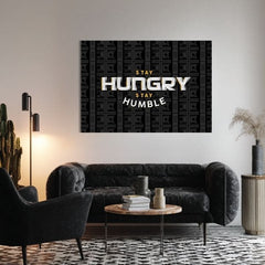 Obraz s motivačním citátem „STAY HUNGRY STAY HUMBLE“ a pozadím bankovek visí v moderním interiéru nad dřevěnou konzolí s vázou a svíčkou Papilora.