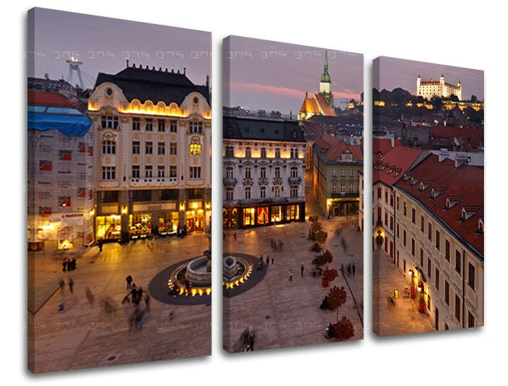 Tři plátna zachycují večerní panorama Bratislavy s osvětlenými budovami a hradem na obzoru, v odstínech fialové a oranžové, Papilora.