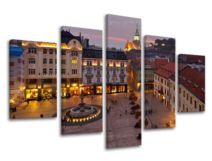 Pohled na osvětlené historické centrum Bratislavy za soumraku, s dominantou kostela a náměstím s fontánou, je zachycen na tomto vícedílném plátně Papilora.