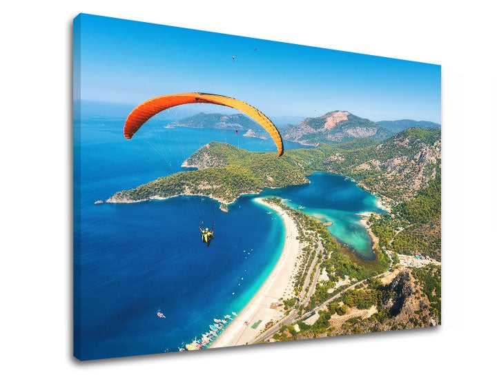 Detailní náhled kompozice zobrazuje paraglidingistu s oranžovým padákem letícího nad zátokou s pláží Papilora.