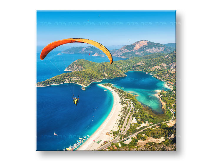 Obraz s paraglidingem nad modrou lagunou a zelenými horami visí v moderním interiéru nad dřevěnou konzolí s vázou a svíčkou Papilora.