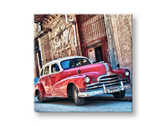 Nástěnná dekorace s motivem červeného retro automobilu v ulicích, zavěšená v moderním interiéru nad dřevěnou konzolí s vázou a svíčkou Papilora.