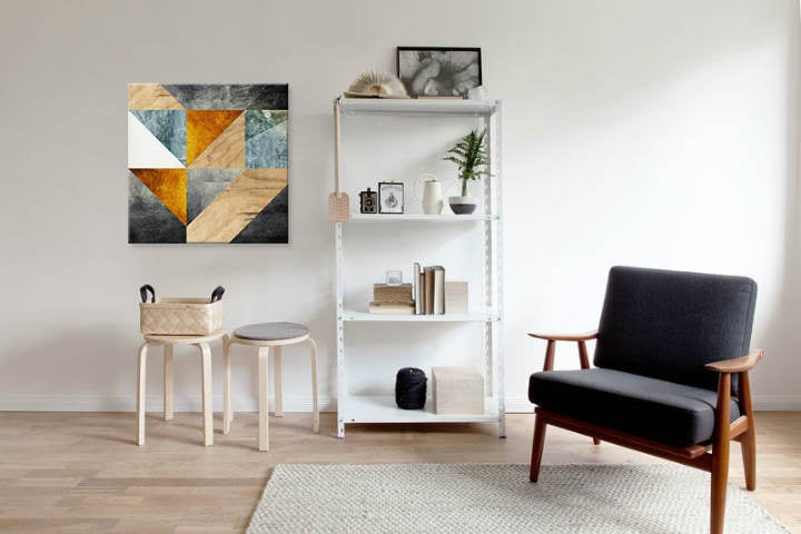 Abstraktní geometrický vzor v šedých, modrých a oranžových tónech visící v moderním interiéru nad dřevěnou konzolí s vázou a svíčkou Papilora.