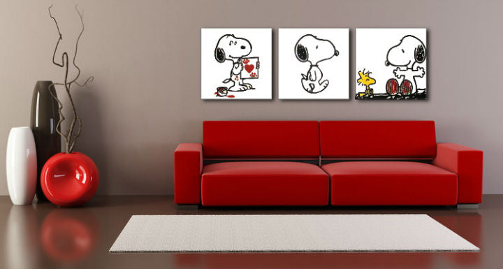 Tři obrazy Snoopyho a Woodstocka v černobílém provedení s červenými detaily visí v moderním interiéru nad dřevěnou konzolí s vázou a svíčkou Papilora.